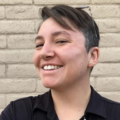 A woman with short brown hair, wearing a black button-down shirt, stands in front of a brick wall. She is smiling, wearing glasses on top of her head.