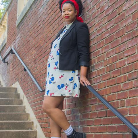 Black woman with red head wrap, natural black hair, wearing a black blazer, a white dress with red and blue flowers, and black boots