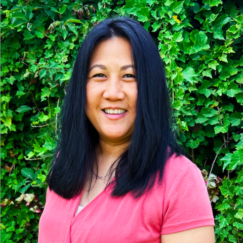 A person with long black hair, wearing a pink top, smiles in front of a leafy green background.