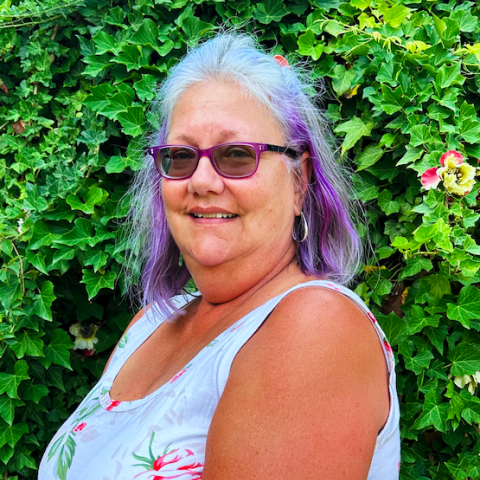 A person with purple-tinted hair and sunglasses smiles while standing in front of a lush green ivy wall. She is wearing a white floral top and hoop earrings, exuding a cheerful and casual vibe.
