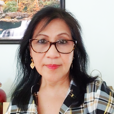 A person with long dark hair and glasses is wearing a plaid shirt and gold earrings. She is seated indoors with a framed scenic picture of a waterfall on the wall behind her.