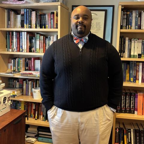 Lamont stands confidently in a room filled with bookshelves. They are wearing a black sweater, a patterned bow tie, and light-colored pants. The shelves are packed with books and a framed certificate is visible on the wall behind them.