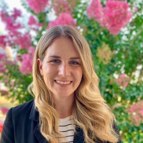 Smiling person with long blonde hair wearing a black jacket and striped shirt, standing in front of vibrant pink flowering trees.