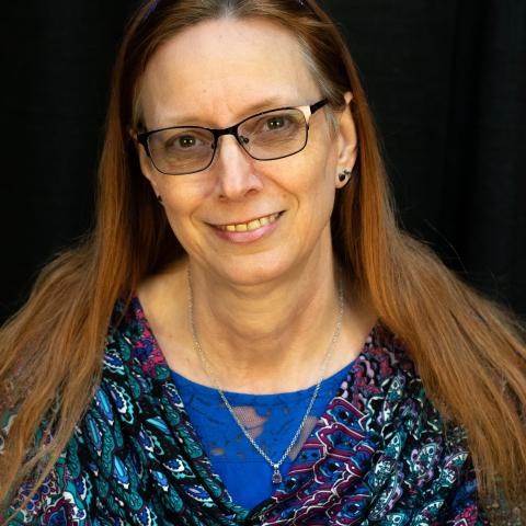 A woman with long auburn hair and glasses, wearing a paisley top with blue, smiles against a dark background.