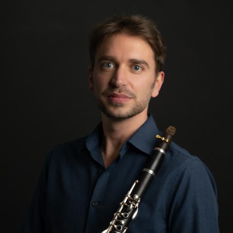 A person with short hair holds a clarinet against a dark background. They are wearing a dark blue shirt and a bracelet, looking at the camera with a neutral expression.
