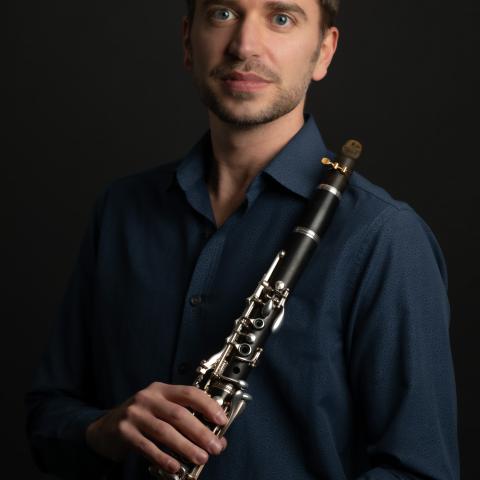 A person with short hair holds a clarinet against a dark background. They are wearing a dark blue shirt and a bracelet, looking at the camera with a neutral expression.