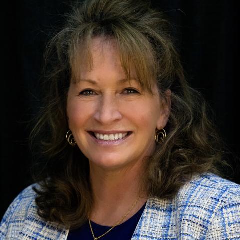 Smiling person with wavy hair wearing a blue and white jacket and a gold necklace against a dark background.