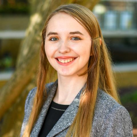 A person with long hair and a grey blazer smiling outdoors with blurred trees in the background. (Kathryn Brinegar)