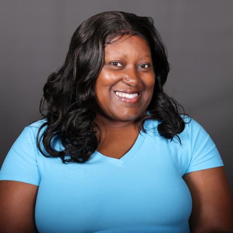 Smiling person with long, wavy black hair wearing a light blue shirt against a plain dark background.