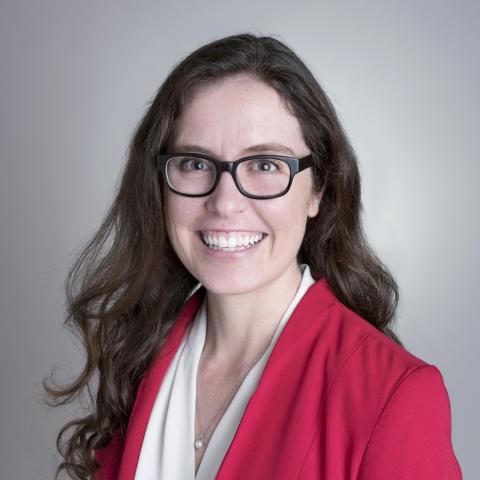 Woman with long brown hair and glasses wearing a red blazer and white blouse smiles at the camera against a plain background.