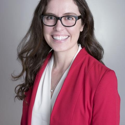 Woman with long brown hair and glasses wearing a red blazer and white blouse smiles at the camera against a plain background.