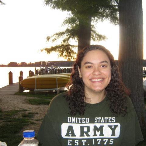 A woman with curly hair smiles at the camera. She is wearing a green "United States Army" t-shirt. Kayaks and boats are visible in the background near a lakeside at sunset.