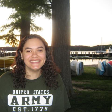 A woman with curly hair smiles at the camera. She is wearing a green "United States Army" t-shirt. Kayaks and boats are visible in the background near a lakeside at sunset.