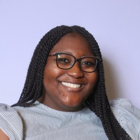 A woman with long braided hair and glasses smiles at the camera, wearing a light striped top, against a plain light purple background.