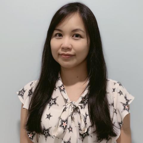 A woman with long black hair is standing against a plain light-colored background, wearing a white blouse with black star patterns and a bow at the collar.