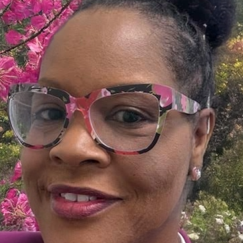 Woman wearing large, colorful eyeglasses and a purple blazer, standing outdoors in front of blooming pink flowers and greenery.