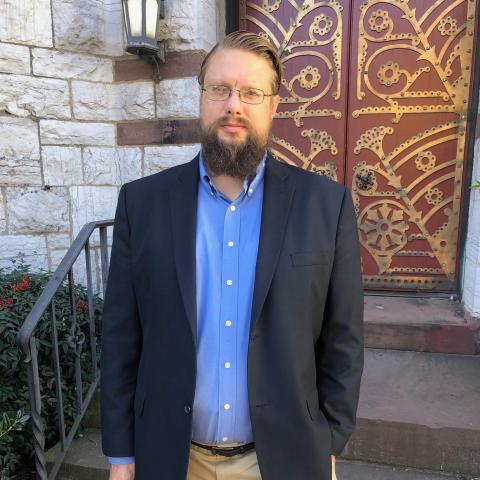 A man with glasses and a beard, wearing a blue shirt and dark blazer, stands outside in front of a decorative red door and stone wall.
