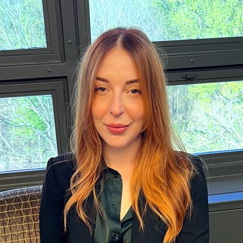 A woman with long red hair and a black blazer sits indoors in front of large windows overlooking green trees.