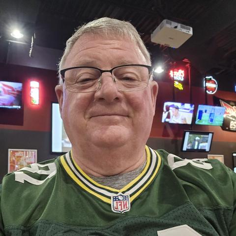Older man wearing glasses and a Green Bay Packers football jersey takes a selfie inside a sports bar with TVs and neon signs in the background.