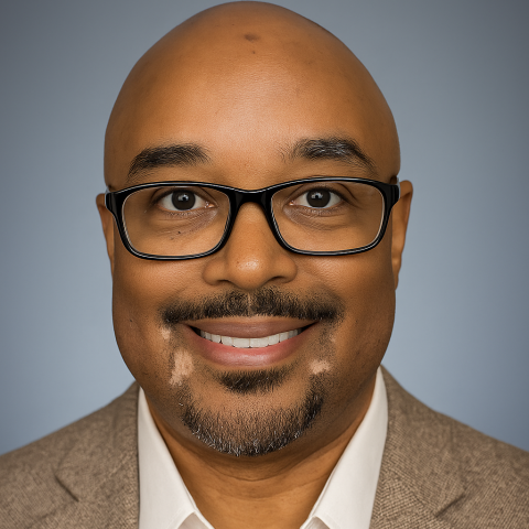 A man with a shaved head, glasses, and a goatee, wearing a tan blazer and white shirt, smiling at the camera against a plain background.