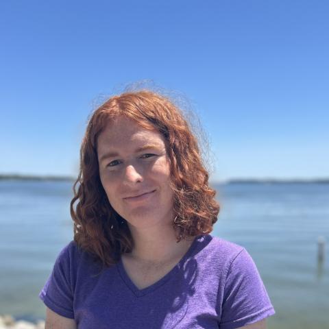A person with curly red hair wearing a purple shirt stands in front of a calm body of water under a clear blue sky.
