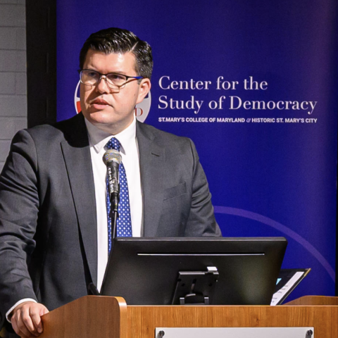 A man in a suit speaks at a podium with a microphone, in front of a banner for the Center for the Study of Democracy at St. Mary’s College of Maryland.