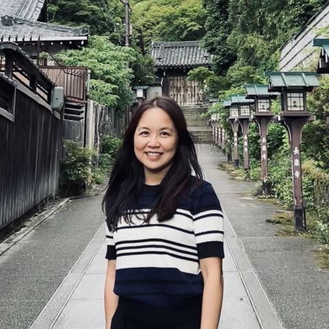 A woman with long dark hair stands smiling on a stone path lined with traditional lanterns and greenery, with wooden buildings and a gate in the background.