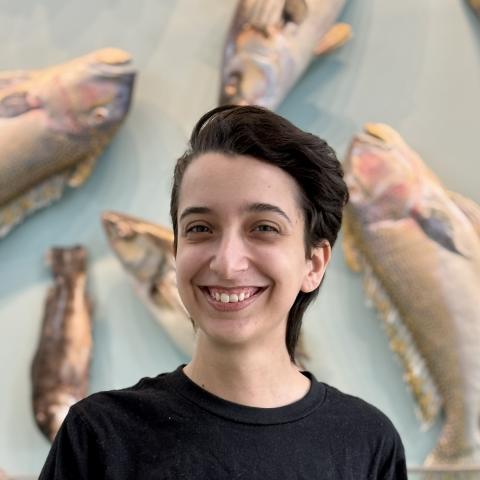 A person with short dark hair and a black shirt smiles in front of a display of mounted fish on a light blue wall.