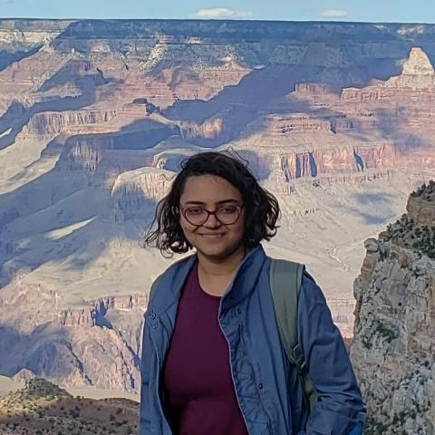 A person with short dark hair and glasses stands in front of the Grand Canyon, wearing a blue jacket and carrying a backpack.