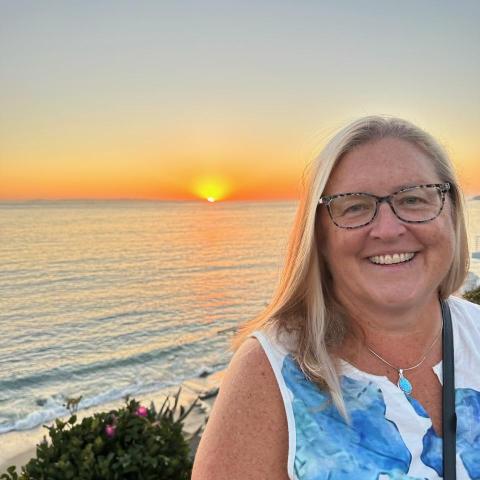 A woman with glasses smiles in front of a beach at sunset, with the sun low on the horizon and calm ocean waves in the background.
