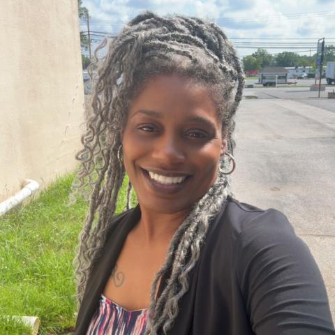 A woman with long, curly gray hair smiles outdoors near a beige building and a parking lot on a sunny day.