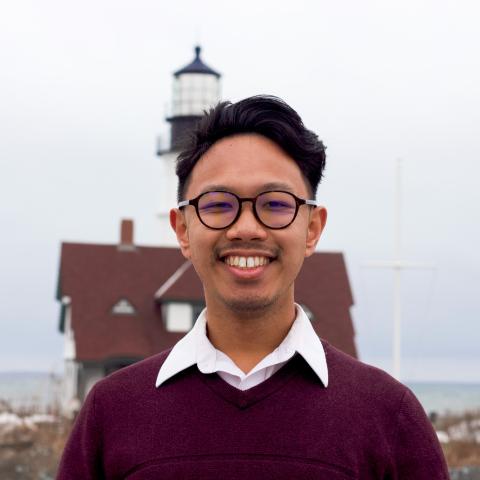 A person wearing glasses and a maroon sweater stands outdoors, smiling, with a house and lighthouse in the background.