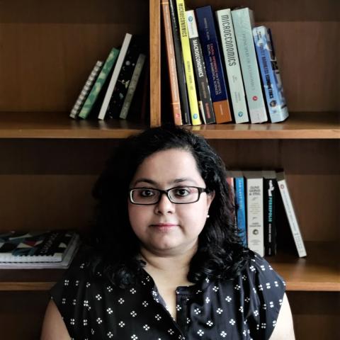 A woman with glasses and dark hair sits in front of a wooden bookshelf filled with books, facing the camera with a neutral expression.