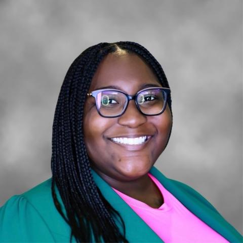 A person with long braided hair, wearing glasses, a green blazer, and a pink top, smiling in front of a gray background.