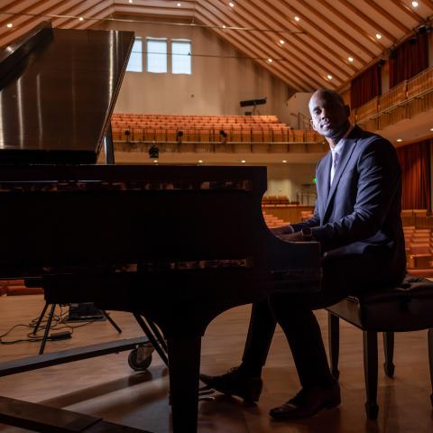 A man in a suit sits at a grand piano on a stage in an empty concert hall, facing the camera.