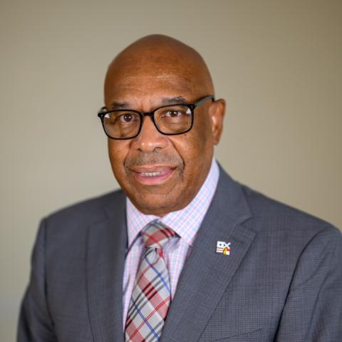 A man wearing glasses and a grey suit with a checkered tie, standing in front of a plain beige background.