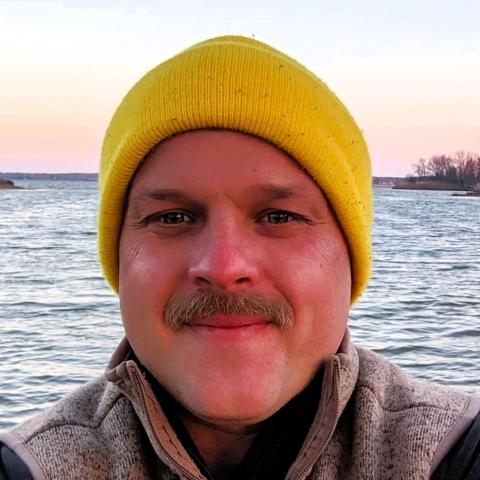 A person wearing a yellow knit hat and gray jacket stands in front of a body of water during sunset.