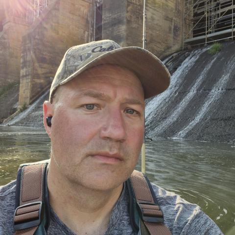 A man wearing a cap in a kayak near a concrete dam with water flowing down its side.