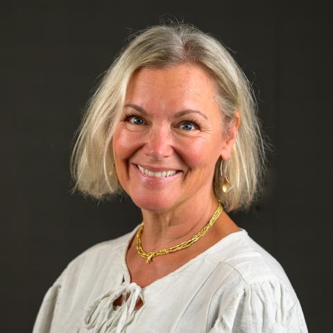 A woman with short blonde hair wearing a white blouse, gold necklace, and earrings smiles in front of a dark background.