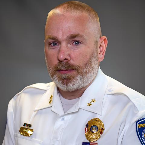 A man with a trimmed beard wearing a white police uniform with badges and shoulder patches poses against a plain, gray background.