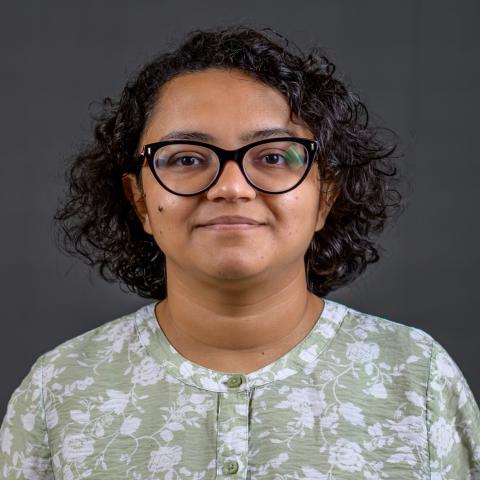 A person with short curly hair and glasses, wearing a light green floral-patterned shirt, stands in front of a plain dark background.