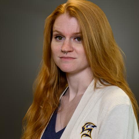 A person with long red hair wearing a cream-colored sweater that has a public safety emblem and text on it, standing against a plain gray background.