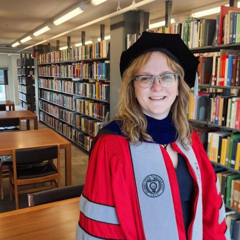 Person in academic regalia stands and smiles in a library, with bookshelves and study tables visible in the background.