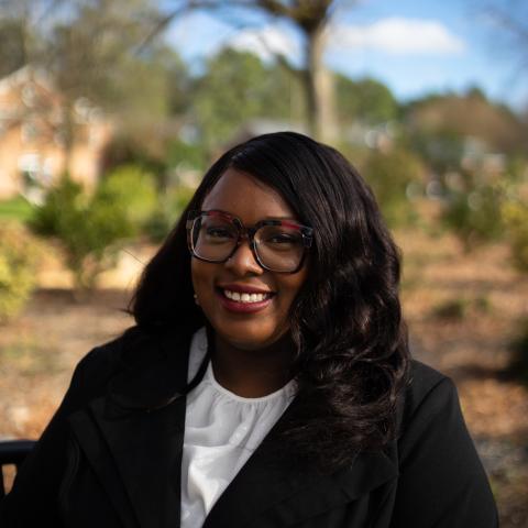 A woman with long dark hair and glasses, wearing a black blazer and white blouse, sits outdoors on a sunny day with trees and bushes in the background.