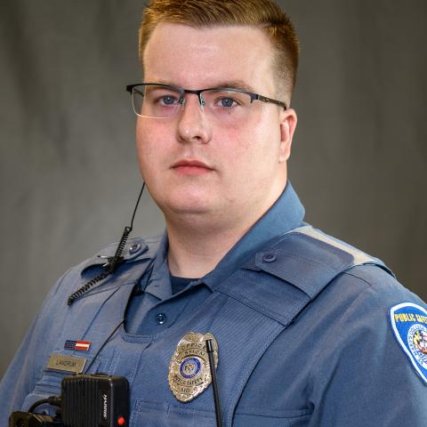  A Public Safety Officer in uniform with a badge, body camera, and radio headset, standing against a plain, neutral background."