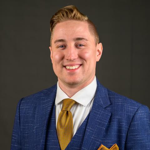 A man wearing a blue suit, white dress shirt, and yellow tie with a matching pocket square stands in front of a plain dark background, smiling at the camera.