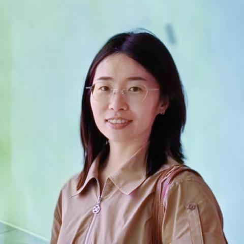 A woman with glasses and shoulder-length dark hair wearing a beige short-sleeve shirt and carrying a bag stands in front of a light green and blue background.