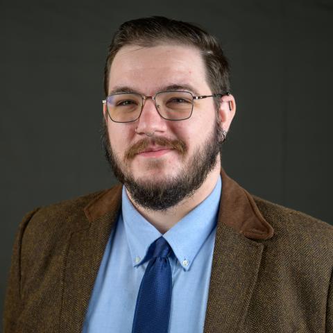 A man with glasses, a beard, and mustache is wearing a brown jacket, blue dress shirt, and blue tie, posing in front of a plain dark background.