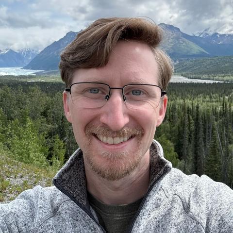 A man with glasses and a light gray jacket smiles at the camera with a forest, mountains, and cloudy sky in the background.