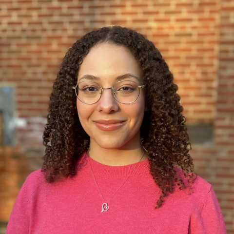 A woman with curly hair and glasses wears a pink top and heart necklace, standing in front of a brick wall.